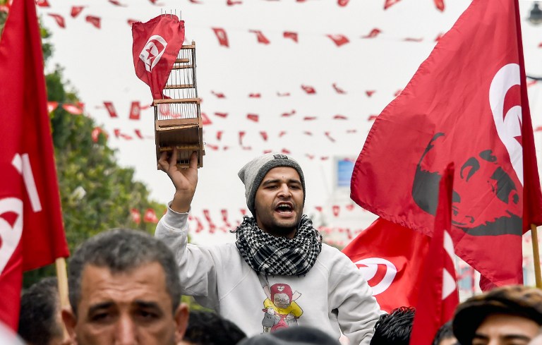 A Tunisian carries a birdcage with the national flag suspended above, next to another national flag defaced with a silhouette of of slain prominent opposition figure Chokri Belaid, during a rally on January 14, 2017 in the Habib Bourguiba Avenue in the capital Tunis to mark the sixth anniversary of the 2011 revolution. / AFP PHOTO / FETHI BELAID